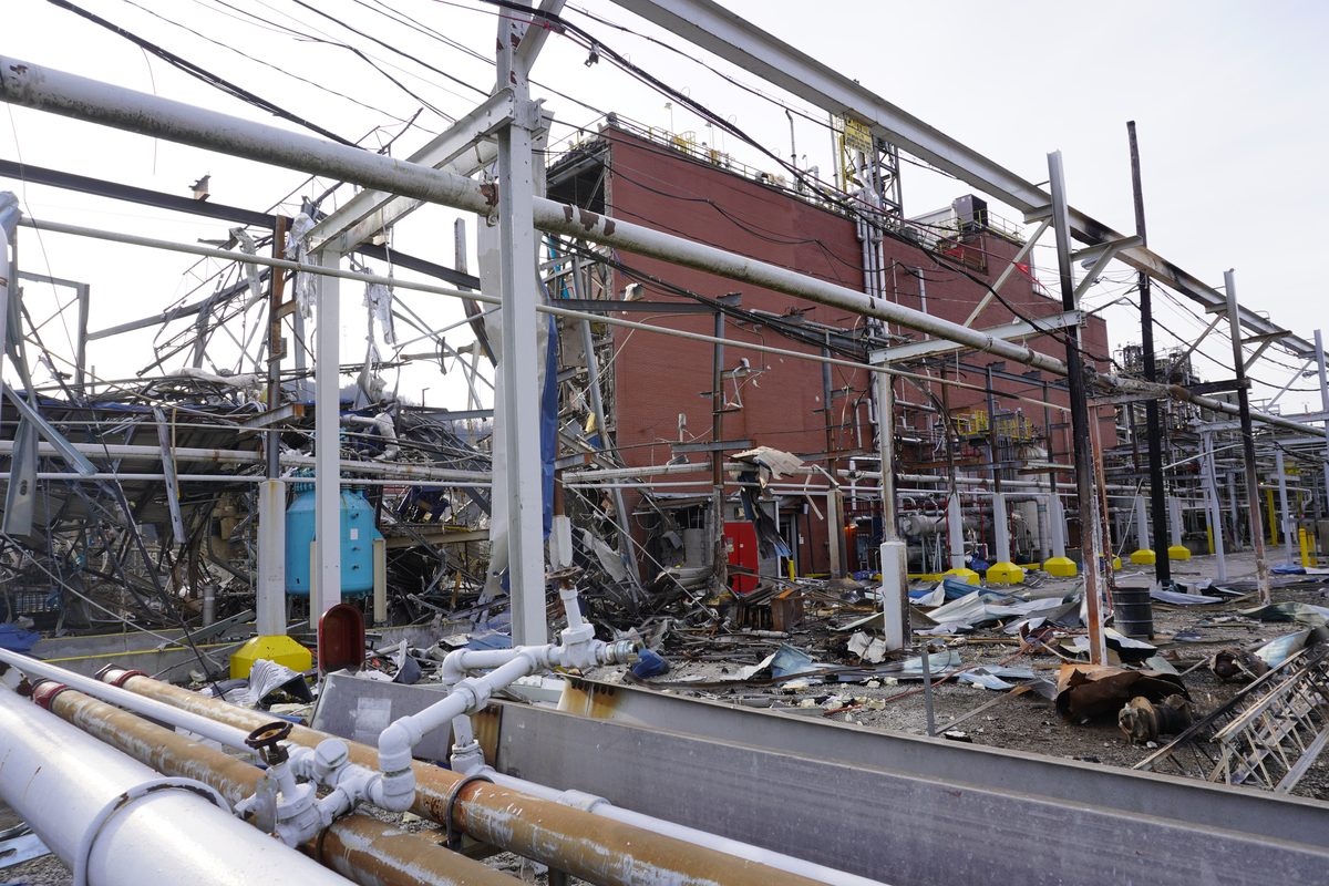 A large industrial building torn open by an explosion inside it, its wall gone and the structural steel and pipework left standing in the debris.