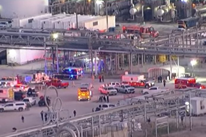 Dozens of fire engines and ambulances assembled across a refinery site at dusk, among pipe racks and process units.