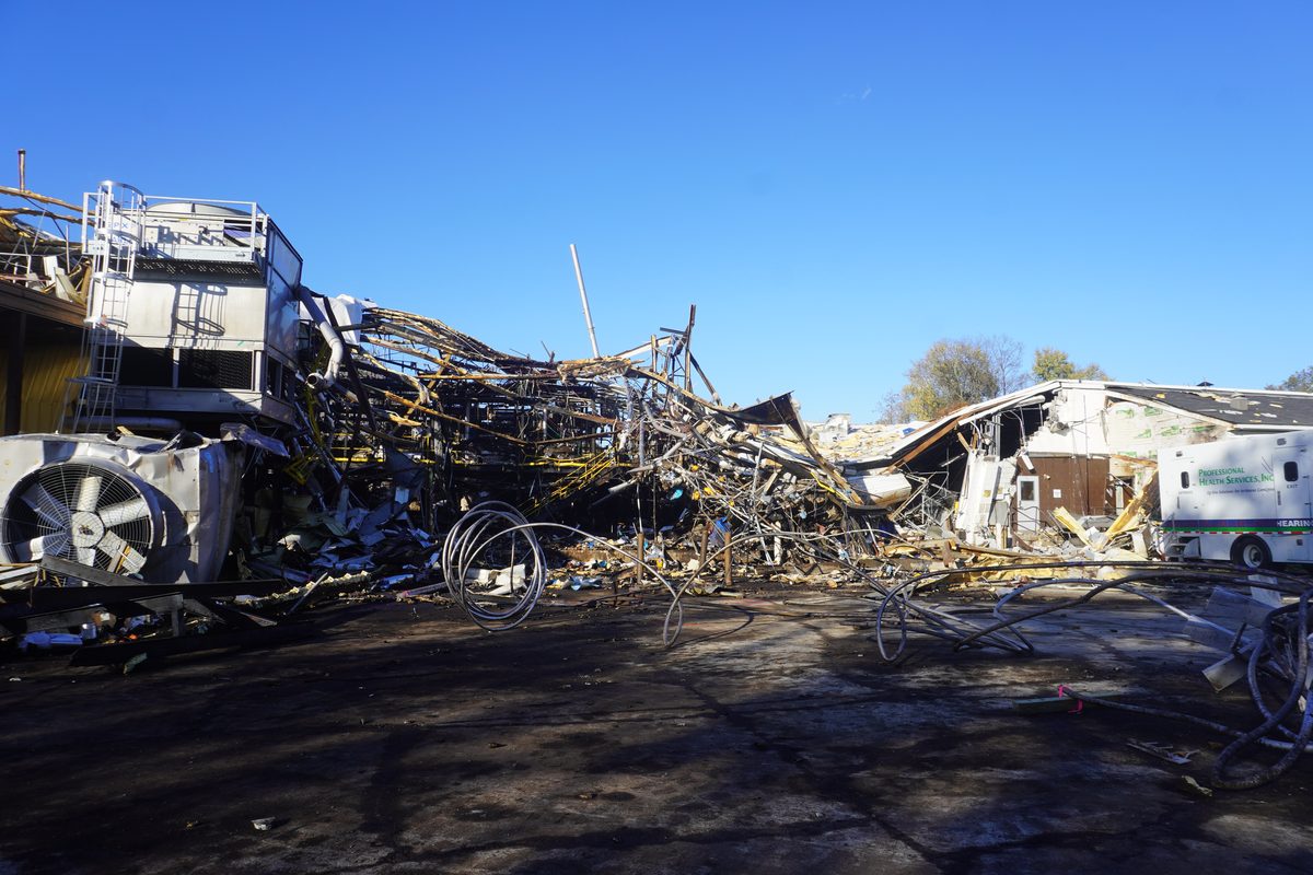 A collapsed industrial building after an explosion, its steel roof structure twisted and fallen across the site.