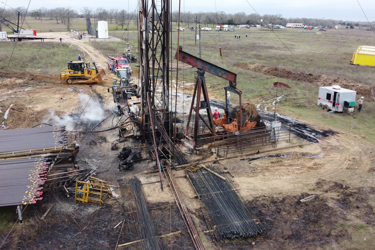 A wellhead site with scorched ground, damaged equipment and emergency vehicles standing off at a distance.